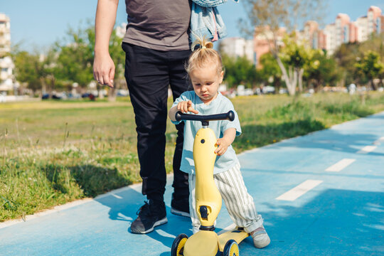 Child learning to ride a yellow scooter in a park with a parent during a sunny day