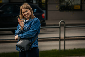 A young woman wearing a denim jacket and crossbody bag waves and smiles at the camera while...