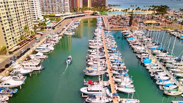 Rows of modern yachts stand at the docks. One boat is moving by the green waterscape. Coast of the Pacific Ocean in Honolulu, Hawaii, USA. Aerial perspective.
