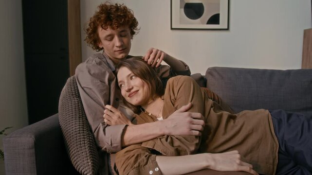 Young man with red curly hair hugging and kissing his girlfriend on head, while they both lying in couch in cozy living rom