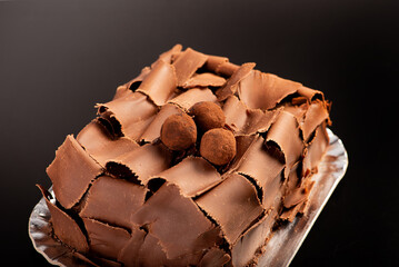 Chocolate cake, close-up of a chocolate cake decorated with rolled chocolate shavings and sprinkled cocoa truffles on a silver tray over a dark surface, selective focus.