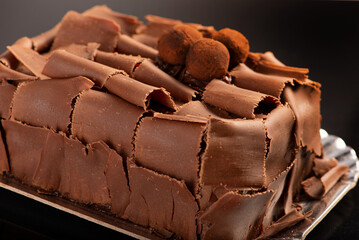Chocolate cake, close-up of a chocolate cake decorated with rolled chocolate shavings and sprinkled cocoa truffles on a silver tray over a dark surface, selective focus.