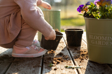 A child in pink shoes and light clothing works with soil in black pots on a wooden deck. A green container with purple and yellow flowers is nearby.