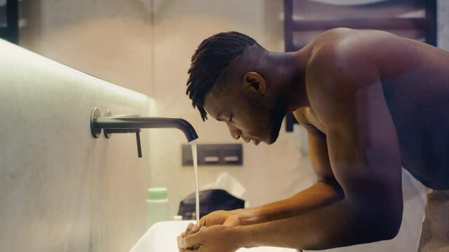 Muscular African American man washing face in bathroom, morning skincare routine
