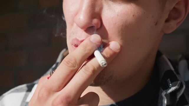 Close up young man in black white checkered flannel shirt holding lighter and cigarette between fingers, puffing smoke stream with brick wall and iron railing blurred in warm sunlight background