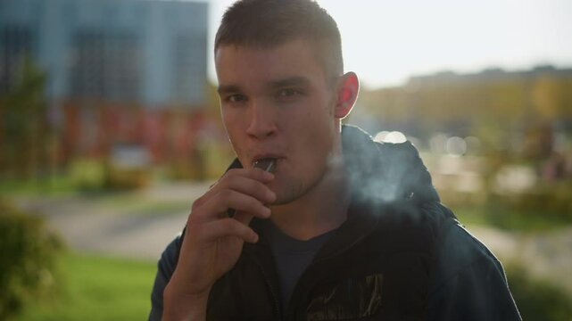 young man in black vest inhales from shisha device and exhales thick white smoke while standing outside in green urban park with modern blurred buildings and evening light in background