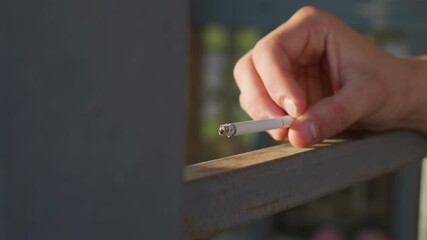 close up of fair skinned man holding lit cigarette over metal railing with wind softly blowing ash while sunlight highlights hand and burning tip during outdoor moment of quiet smoking