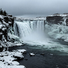 Fototapeta premium Powerful waterfall plunges into a dark, turbulent river surrounded by snow and ice under a gray sky