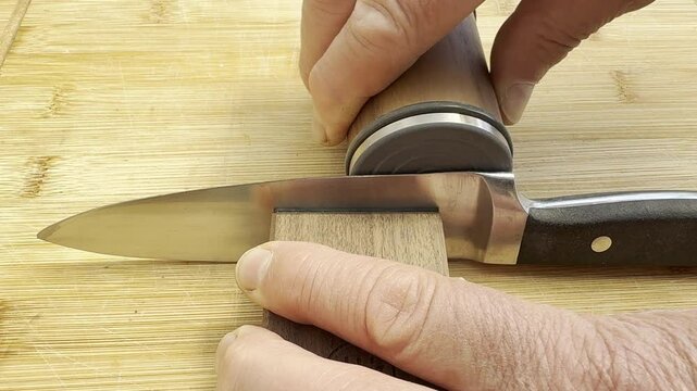 Closeup of hands using a modern knife sharpener, consisting of a magnetic wooden block to hold the knife, while a disc is rolled along the blade length to sharpen it.