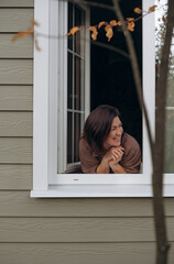 A woman leans out of a window in her cozy home, smiling as she enjoys a moment of tranquility. The...