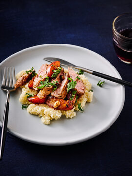 A plate holds a generous serving of fluffy couscous topped with tender beef slices, grilled peach, and a sprinkle of mint. A glass of red wine accompanies the dish, enhancing the experience