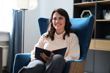 Obraz premium A woman is sitting in a blue chair with a book in her lap. She is smiling and she is enjoying her time reading. Concept of relaxation and leisure