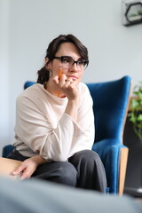 A woman sits in a blue chair with a pencil in her hand. She is looking at something and she is deep...