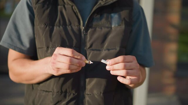 partial view of man in dark vest breaking cigarette into two pieces with both hands outdoors during sunny day, making clear decision to quit smoking, with blurred background