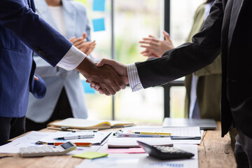 Business partners shaking hands at a meeting while their coworkers are clapping, celebrating a successful agreement.