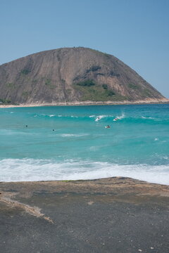 Praia de Itacoatiara, em Niter&oacute;i, Rio de Janeiro, em um dia de ver&atilde;o. Ondas, mar azul, surfista na &aacute;gua e o Cost&atilde;o de Itacoatiara ao fundo sob c&eacute;u limpo.