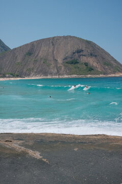Praia de Itacoatiara, em Niter&oacute;i, Rio de Janeiro, em um dia de ver&atilde;o. Ondas, mar azul, surfista na &aacute;gua e o Cost&atilde;o de Itacoatiara ao fundo sob c&eacute;u limpo.