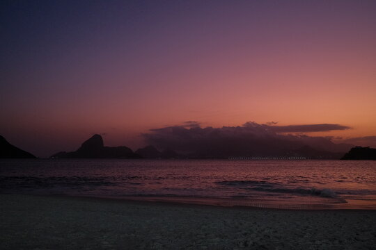 P&ocirc;r do sol vibrante na Praia de Icara&iacute;, em Niter&oacute;i, com vista para o P&atilde;o de A&ccedil;&uacute;car, Ba&iacute;a de Guanabara e o skyline do Rio de Janeiro sob tons de rosa, roxo e laranja.