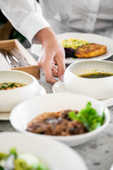 A person's hand reaches for a bowl of soup amidst a selection of beautifully presented dishes. The setting showcases a variety of colorful meals in a bright dining atmosphere.