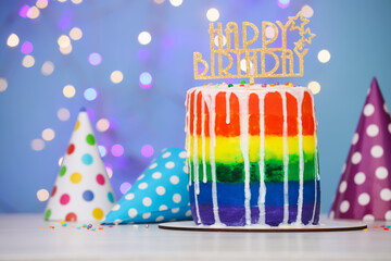 Delicious rainbow birthday cake with topper and party hats on white table against light blue...