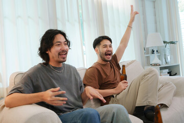 Fototapeta premium Two Asian Male friends are having a fun game night in a living room, sharing drinks and cheering excitedly as they play together.