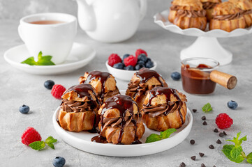 Profiteroles with chocolate cream, fresh berries and chocolate glazed on a plate on a gray concrete background. A delicious French dessert.