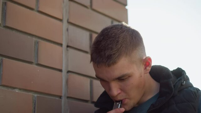 white boy leaning against brick wall outdoors inhaling from vape device into mouth as he coughs while examining device wearing black puffer vest over dark shirt