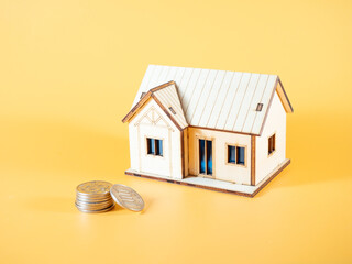 A small model house with a blue roof and windows sits on a yellow background, next to a stack of coins
