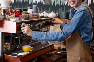 Asian young woman operating espresso machine at coffee shop. Wearing brown apron and denim shirt while making fresh drink. Smiling expression reflects joy in small cafe business.