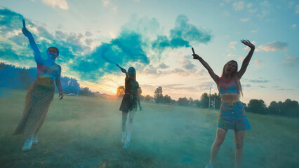 Three young women, likely school friends, celebrate with blue smoke bombs in a field at sunset,...