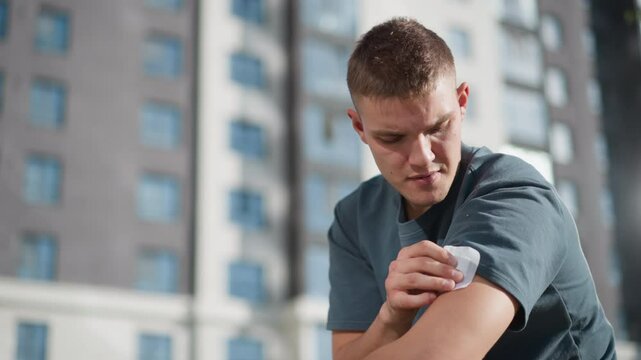 young man lifting sleeve to apply nicotine patch on upper arm while sitting outdoors with blurry view of high-rise residential building in background