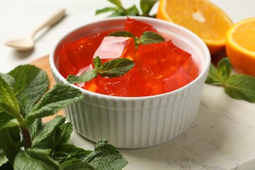 Delicious jelly cubes in bowl, mint, spoon and orange on table, closeup