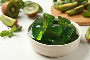 Delicious green jelly cubes in bowl, mint and kiwi on white table, closeup