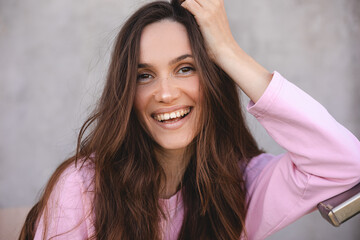 Portrait of beautiful brunette long hair woman wear pink long sleeve shirt, hold head and standing near stairs. Girl laughing and smiling, positive emotions.