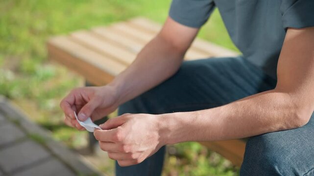 partial view of young man seated on wooden bench outdoors holding and unwrapping nicotine patch under sunlight with green grass and pavement in background casual clothing and focused gesture visible