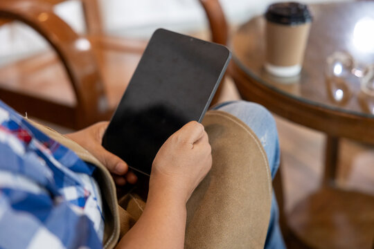 Closeup of senior woman in brown apron sitting in cafe holding tablet. Casual moment captures digital technology use in small business, relaxing with coffee cup on table nearby.