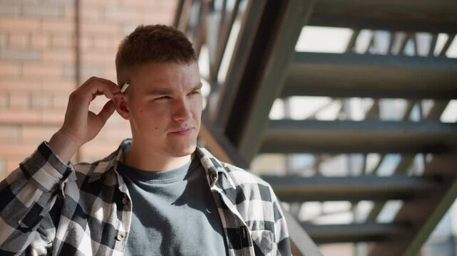 portrait view of student in black white plaid shirt on metal staircase returning cigarette to ear while looking at camera against sunlit brick wall and blurred staircase under midday sun