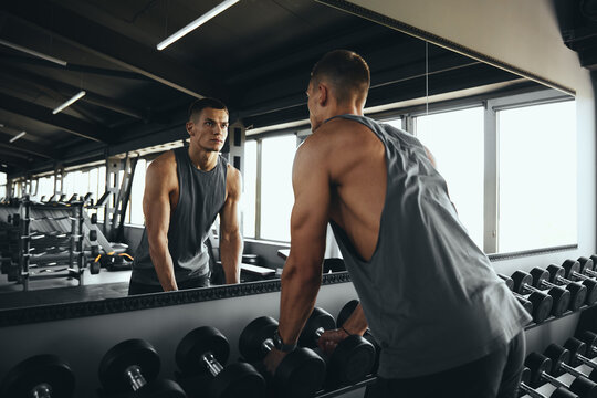 Horizontal photo of attractive bodybuilder man in sportswear looking in mirror while choosing weights at modern gym for workout. Bodybuilding, fitness and health care concept.