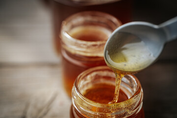 A close-up of golden honey slowly dripping from a plastic spoon into a glass jar, with more jars in the background softly blurred on a rustic wooden surface.