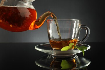 Pouring freshly brewed tea from teapot into glass cup on black table, closeup