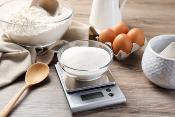 Digital kitchen scale with bowl of sugar among other products on wooden table