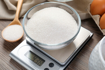 Digital kitchen scale with bowl of sugar on wooden table, closeup