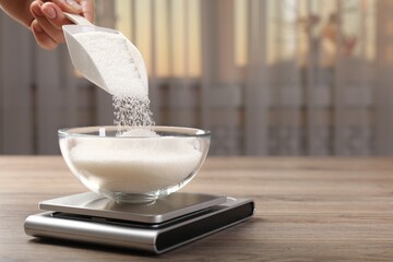 Woman adding sugar into bowl and digital kitchen scale at wooden table indoors, closeup. Space for...