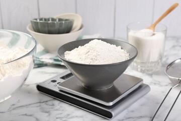 Digital kitchen scale with bowl of flour and other products on white marble table, closeup