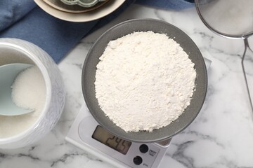 Digital kitchen scale with bowl of flour and other products on white marble table, flat lay