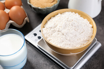 Digital kitchen scale with bowl of flour and other products on grey table, closeup
