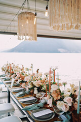Long festive table with bouquets of flowers stands on the pier under a canopy with macrame lamps