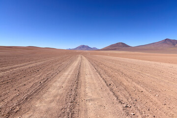 Dirt road stretches toward distant volcanic mountains across vast empty plain in Eduardo Avaroa National Park, Bolivia. Perfect blue sky over Andean high desert.
