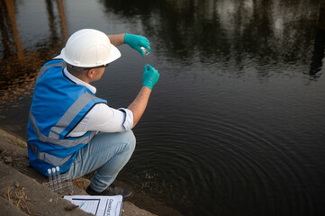 A water resource worker is bending down to collect water samples from a canal into test tubes for...