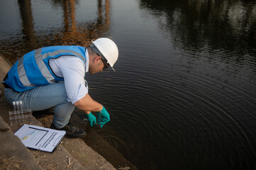 A water resource worker is bending down to collect water samples from a canal into test tubes for...